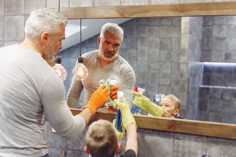 Small boy helping grey haired dad with beard cleaning mirror in tile bathroom